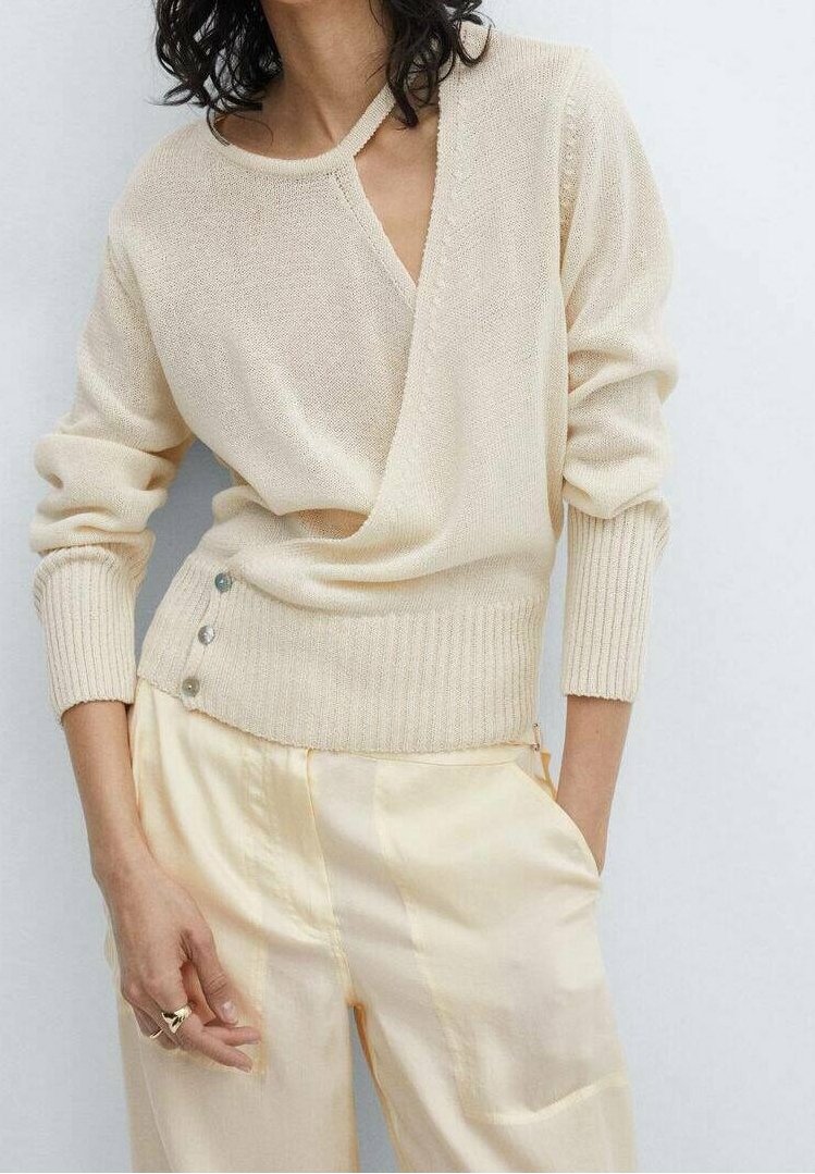 Femme portant un pull en maille crème avec des boutons sur le côté et un pantalon en satin crème assorti, debout, une main dans la poche.