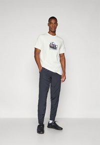 T-shirt beige clair à manches courtes avec un motif graphique sur le devant, associé à un pantalon gris foncé et des baskets noires. Tenue simple et décontractée.