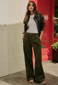 Jachetă din piele neagră peste o cămașă albă, asortată cu pantaloni largi din material tartan verde măsliniu și pantofi bej, plasată în fața unui fundal cu ușă roșie.
