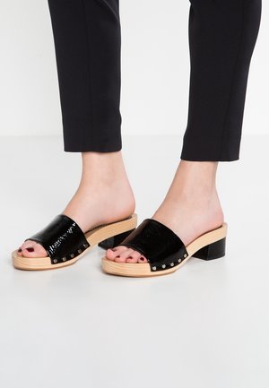 Fødder iført sorte slip-in sandaler i laklæder med chunky træsåler, kombineret med sorte smalle bukser på en hvid gulv.