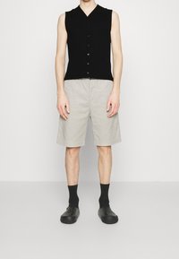 Person trägt eine ärmellose schwarze Knopfweste, beigefarbene, knielange Shorts, schwarze Socken und schwarze Slipper und steht vor einem schlichten Hintergrund.