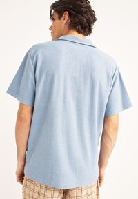 Camiseta polo de manga corta en azul claro, hecha de una tela texturizada. Presenta un cuello plano y un corte relajado, con una etiqueta visible en el lado.