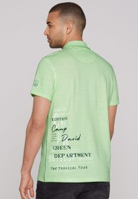 Zelené pruhované polo tričko s golierom, so čiernymi textovými grafikami, vo voľnom strihu, s krátkymi rukávmi a hladkou textúrou látky.
