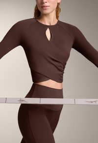 Vrouw draagt een nauwsluitende bruine cropped top met lange mouwen en een legging met een beige elastische band met kangoeroe-silhouetten rond haar taille.