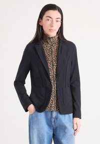 Schwarzer Blazer mit zwei Fronttaschen und Reverskragen, getragen über einem braunen Leopardenmuster-Hochkragen-Top. Bequeme blaue Jeans runden den Look ab.