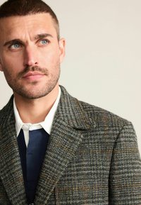 Grå tweedfrakke med et ternet mønster, der har en udskåret krave, tekstureret stof og en lagdelt navy- og hvid skjorte under.