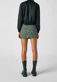 Černá bomber bunda, zelená strukturovaná mini sukně se zigzag vzorem a černé vysoké bojové boty. Hladké materiály a přiléhavý design.