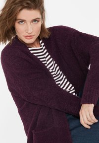 Frau in einem dunkellila strukturierten Cardigan über einem schwarz-weiß gestreiften Hemd, sitzend mit überschlagenen Händen auf dem Schoß.