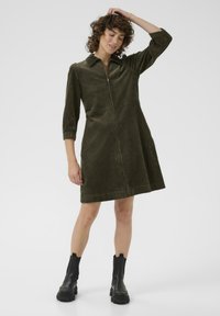 Abito in corduroy verde oliva con zip frontale, colletto e maniche a tre quarti. Abbinato a stivali neri alla caviglia.