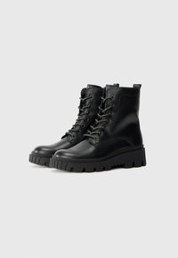 Botas tobilleras negras de cuero con suela de goma. Entre sus características se incluyen punta redonda, cordones en la parte delantera y una lengüeta en la parte trasera. Detalles texturizados.