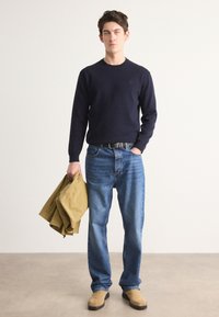 Navy knit sweater, blue denim jeans, beige light jacket held in hand, and tan shoes; simple design with minimalistic branding.