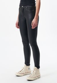 Černé skinny džíny ve vzhledu kůže s vysokým pasem, obsahující přední kapsy, zip a stříbrné detaily, doplněné béžovými vysokými teniskami.