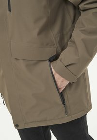 Olivgrön ytterjacka med en texturerad finish, med svarta dragkedja detaljer och en framlapp; figursydd design med en stilren look.