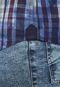 Kurzärmliges kariertes Hemd in Blau-, Rot- und Grüntönen mit einem dunkelblauen Saum, kombiniert mit hellen Jeans in Denim-Optik, die eine strukturierte Oberfläche zeigen.