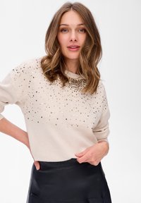 Beżowy sweter z cekinami ułożonymi w gradientowy wzór, okrągłym dekoltem i długimi rękawami. Połączony z czarną skórzaną spódnicą.
