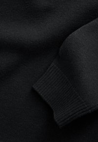 Pull en maille noire montrant des poignets côtelés et une texture lisse. Le tissu semble doux et extensible avec un motif uniforme partout.