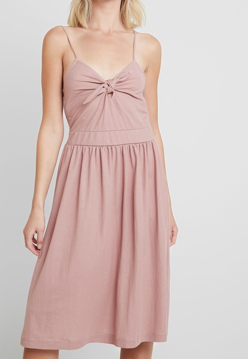 Robe rose sans manches avec un détail noué à l'avant, un haut ajusté et une jupe évasée. Fabriquée en tissu doux et léger.