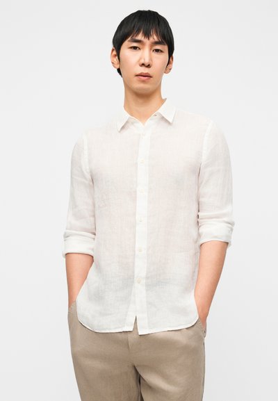 Λευκό πουκάμισο από λινό με κουμπιά, μανίκια γυρισμένα, κλασικό γιακά και διακριτική υφή. Συνδυασμένο με ανοιχτό μπεζ παντελόνι.