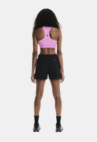 Top corto atlético rosa, pantalones cortos negros con cintura elástica, calcetines negros y zapatillas deportivas. Pelo rizado, de pie con la espalda hacia la cámara.