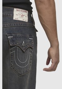 Ciemnoniebieskie dżinsy z markowym metkiem na pasku, z białymi szwami i charakterystycznym wzorem kieszeni w formie zakrzywionej linii.