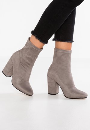 Bottines en daim taupe à talons carrés portées avec des jeans skinny noirs à ourlets effilochés sur un sol blanc lisse.