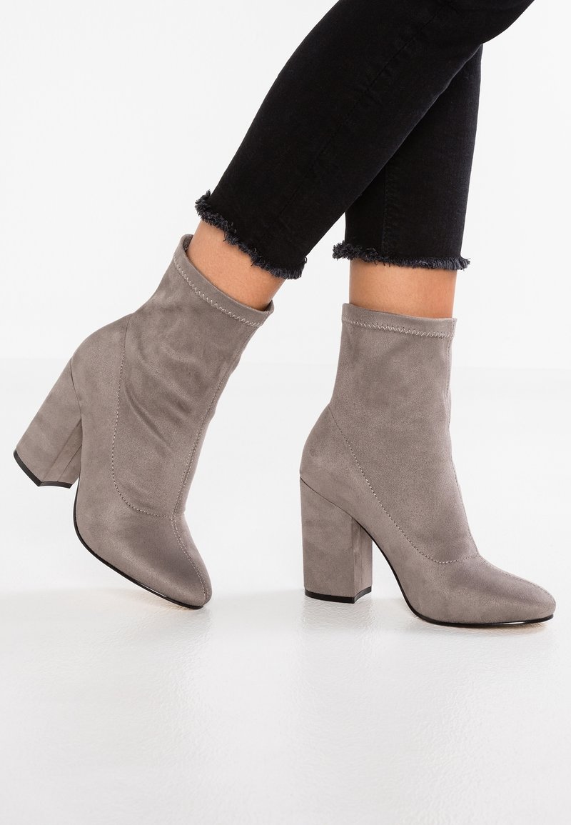 Bottines en daim taupe à talons carrés portées avec des jeans skinny noirs à ourlets effilochés sur un sol blanc lisse.