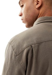 Krave skjorte i afdæmpet beige stof med en struktureret overflade og synlige syninger, vist fra en trekvart bagvinkel.
