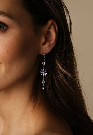 Close-up van het oor van een vrouw met een lange zilveren oorbel met paarse hartvormige en bloemvormige edelstenen.