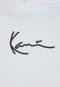 Tissu blanc avec du texte noir brodé qui ressemble à une signature manuscrite. Le design est lisse avec une texture fine.