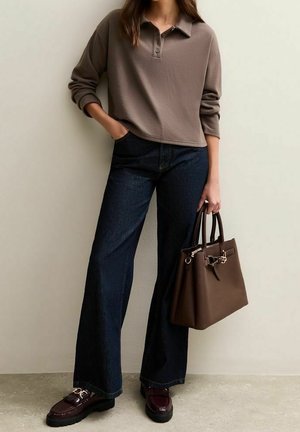 Femme portant un polo marron à manches longues, un jean large foncé, des mocassins marron, tenant un grand sac à main marron contre un mur clair uni.