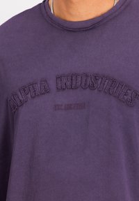 Fialová bavlnená tričko s vyšitým textom "ALPHA INDUSTRIES" a "EST. SINCE 1959", s okrúhlym výstrihom a jemne obnošeným lemom.