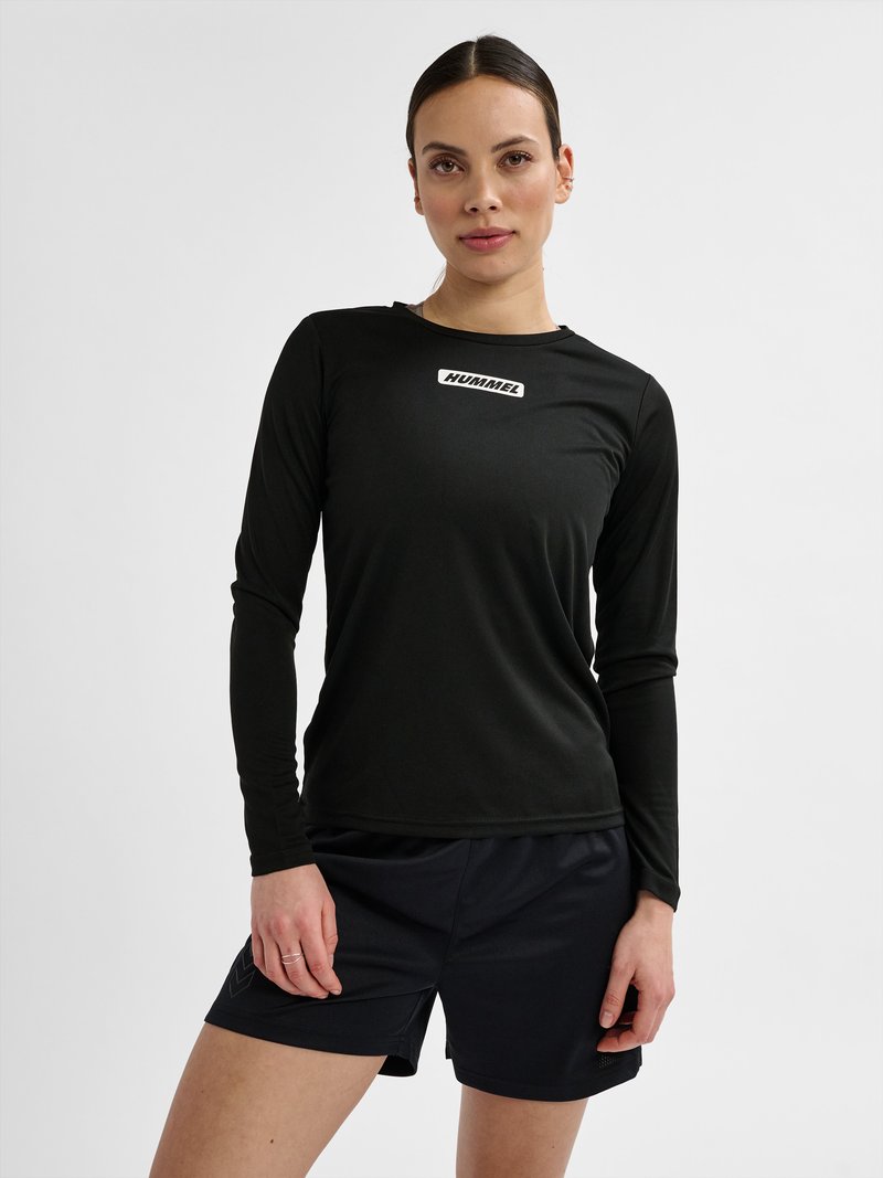 Femme aux cheveux attachés, portant un t-shirt Hummel noir à manches longues et un short noir, se tenant devant un fond blanc uni.