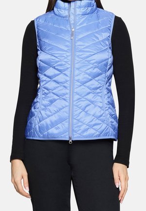Femme portant une doudoune sans manches matelassée bleu clair avec une fermeture éclair devant, par-dessus un haut à manches longues noir et un pantalon noir.