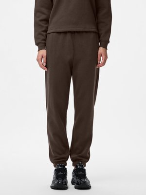 Personne debout portant un pantalon de survêtement marron foncé, un sweat assorti et des chaussures de sport noires, visible des épaules vers le bas sur fond uni.