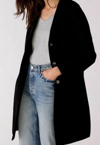 Svart ullblandad kappa med slagkrage, prydd med två stora knappar, bärs över en grå t-shirt med v-ringning och blå jeans med hög midja.