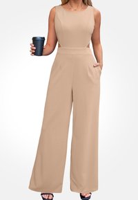 Salopette beige senza maniche con design a gamba larga, ritagli laterali e tasche frontali. Modella tiene in mano una tazza nera, indossando una collana semplice.