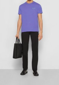 Lila bomullströja med grön text, i kombination med svarta slim-fit byxor och svarta lädersneakers. En svart tote-väska hålls i en hand.