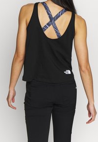 Femme portant un haut sans manches noir sur un soutien-gorge de sport à dos croisé à motifs avec un pantalon noir, se tenant de dos face à l'appareil photo.