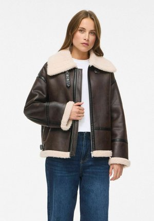 Une femme porte une veste en peau lainée marron foncé avec un col et des poignets crème, une chemise blanche et un jean bleu sur un fond uni.