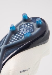 Acercamiento a la suela blanca de un botín de fútbol Adidas con tacos azules y textura de agarre, destacando la tecnología SprintFrame y el diseño Agility Bandage.