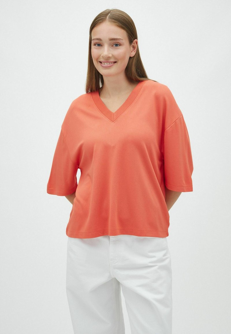Jeune femme aux cheveux bruns lisses portant un haut ample corail à col en V et manches courtes ainsi qu'un pantalon blanc sur un fond clair uni.