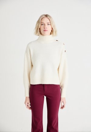 Suéter color crema con un alto cuello acanalado, que presenta un detalle de botón en el hombro, combinado con pantalones ajustados color burdeos.
