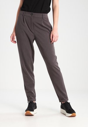 Vrouw met donkergrijze taps toelopende broek met omgeslagen boorden en zwarte platformsneakers, staand tegen een effen lichte achtergrond.