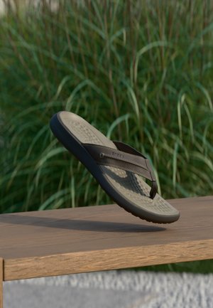 Brune og beige Crocs flip-flops hængende over en træbænk med grøn græs i baggrunden.