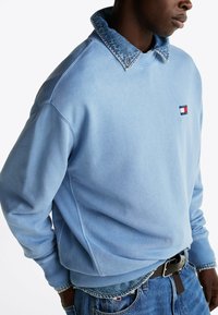 Jasnoniebieski sweatshirt z denimowym kołnierzem, prążkowanymi mankietami i naszywką z logo na piersi. Posiada luźny krój i miękką teksturę.