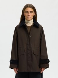 Kvinde med langt brunt hår iført en mørkebrun jakke med sort krave og manchetdetaljer over en beige rullekravesweater.