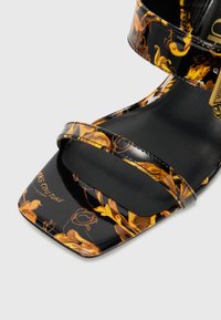 Sandal em pele de patente preta com padrões florais dourados, duas correias finas, bico quadrado e palmilha texturizada preta com logo.