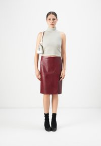 Žena v bezrukávovom béžovom pruhovanom roláku, bordovej koženej sukni, čiernych členkových čižmách a s bielou ramennou taškou na bielom pozadí.