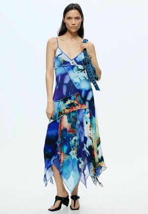 Naine kannab voolavat sinist abstraktse mustriga kleiti, millel on õhukesed rihmad, hoides sinist käekotti ja kandes musti kontsaga sandaale.