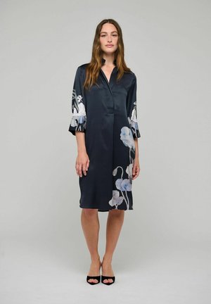 Donna in piedi che indossa un vestito scuro al ginocchio con motivi floreali chiari sulle maniche e sull'orlo, abbinato a tacchi neri open toe.